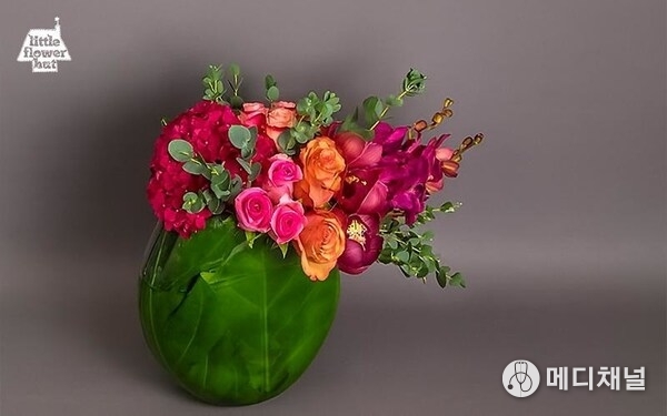 Vibrant floral arrangement in a glass vase wrapped with green leaves, featuring roses, hydrangeas, and orchids.